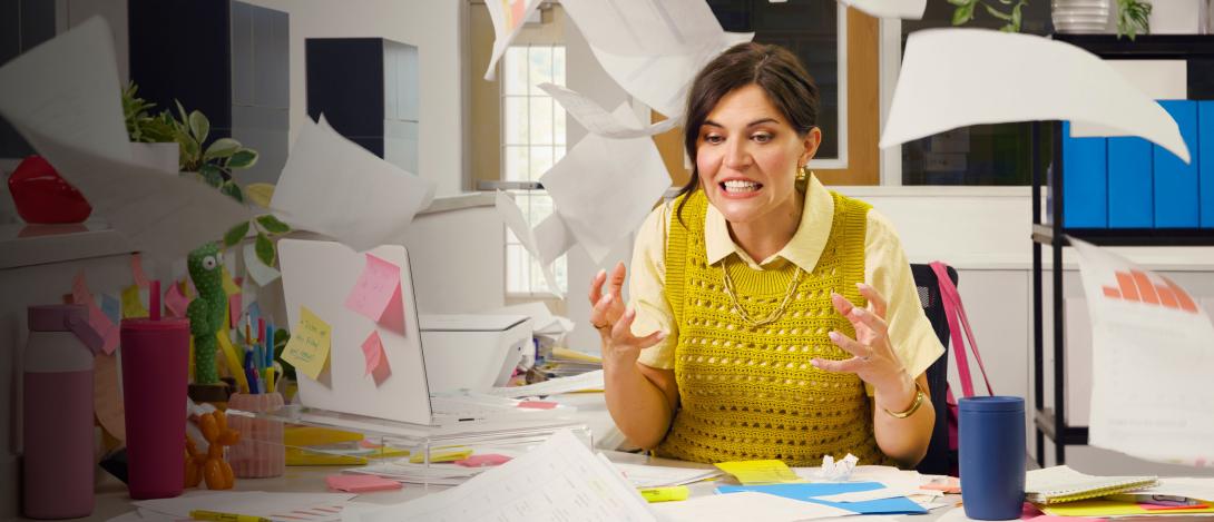 A woman grits her teeth in frustration as office papers fly around her desk.