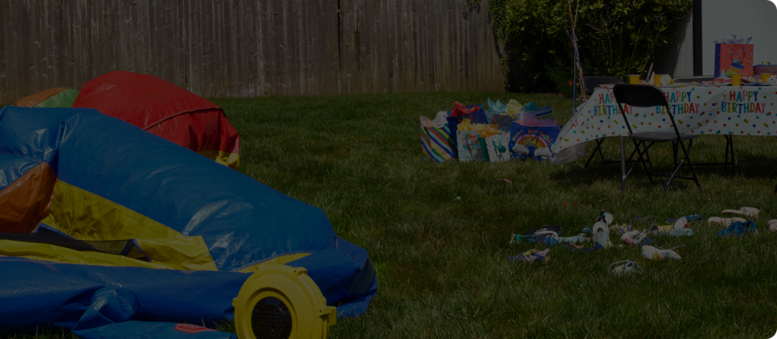 A chaotic birthday party scene, with an uninflated bouncy castle and party debris on the grass.