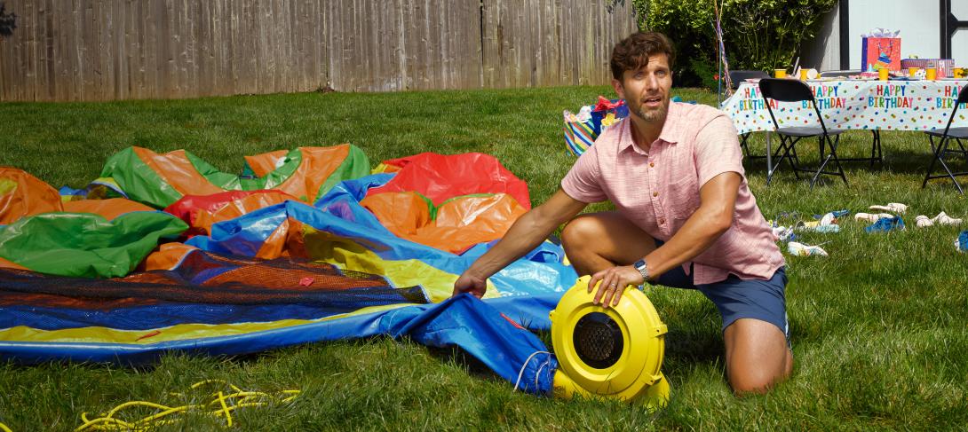 A man has a panicked expression while struggling to inflate a bouncy house.