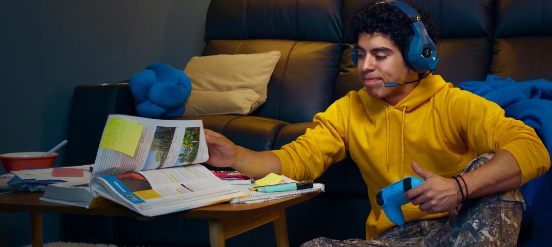 A teenager pages through a textbook while holding a video game controller.