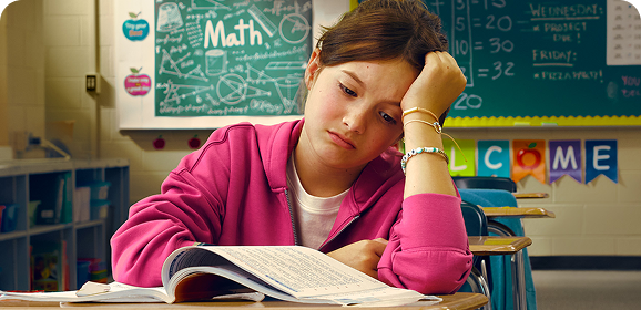 An elementary-aged girl stares in frustration at a textbook, head in hand.