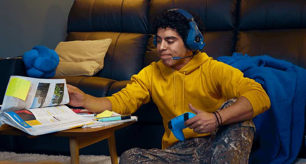 A teenager pages through a textbook while holding a video game controller.