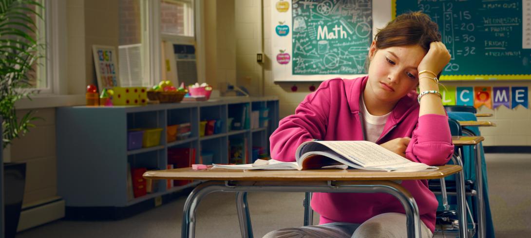 An elementary-aged girl stares in frustration at a textbook, head in hand.